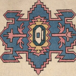 K&ouml;nnte beinhalten: Ein Vintage-Teppich mit einem geometrischen Muster in Rosa-, Blau- und Gelbt&ouml;nen. Der Teppich hat einen wei&szlig;en Hintergrund und ein zentrales Design mit einer Diamantform.