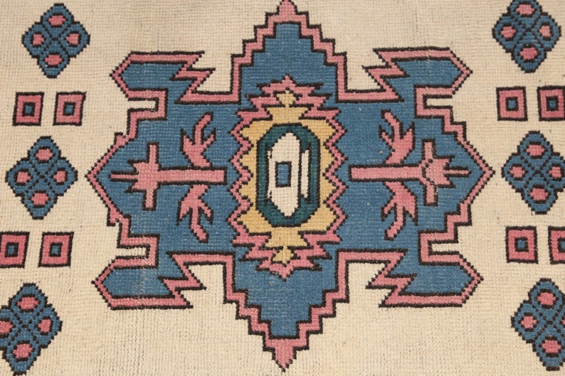 K&ouml;nnte beinhalten: Ein Vintage-Teppich mit einem geometrischen Muster in Rosa-, Blau- und Gelbt&ouml;nen. Der Teppich hat einen wei&szlig;en Hintergrund und ein zentrales Design mit einer Diamantform.
