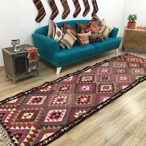 May include: A long rectangular runner rug featuring a repeating geometric diamond pattern in shades of pink, orange, brown, and cream. The rug has a dark border and light-colored fringe at both ends, showcasing a woven texture throughout the piece.
