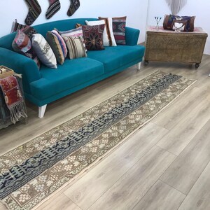 May include: A long, rectangular runner rug featuring a central dark blue geometric pattern flanked by light brown borders with repeating floral motifs. The rug displays a weathered texture with intricate woven details throughout its length.