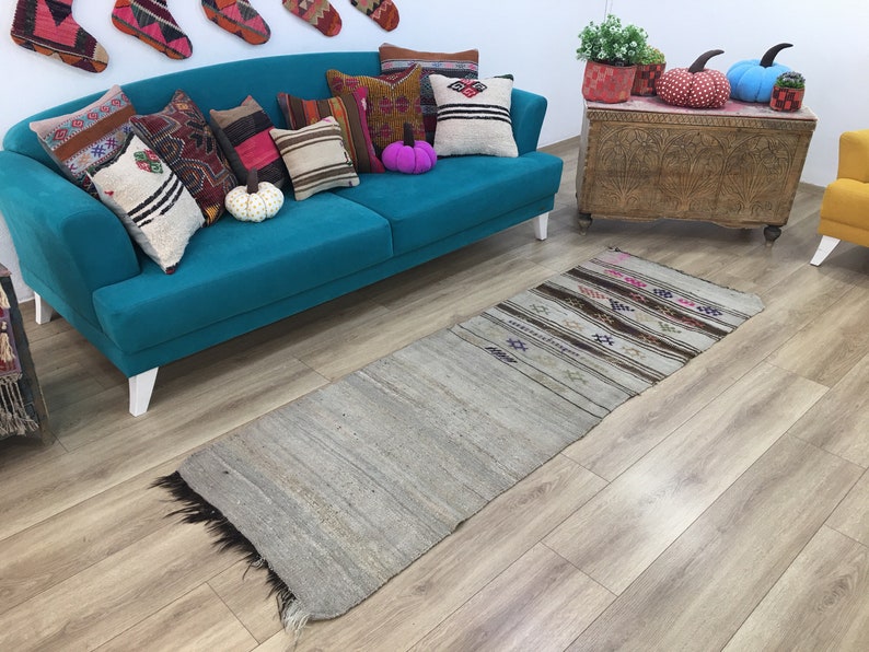 May include: A teal blue sofa with white legs, decorated with colorful throw pillows and a patterned rug. The sofa is in a room with a wooden chest and a yellow chair.