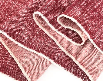 May include: A deep red textile with a textured, woven appearance featuring a lighter pinkish-red underside. The fabric displays a consistent grid-like pattern of white flecks throughout the surface, with finished edges visible along the folded sections.