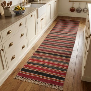 May include: A long rectangular runner rug featuring horizontal stripes in shades of red, black, yellow, and white. The rug has a woven texture and is finished with short, light-colored fringe tassels along both of the shorter ends of the textile.