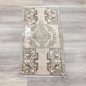 May include: A rectangular area rug featuring a central geometric medallion pattern in shades of beige, light brown, and muted sage green. The design includes symmetrical angular motifs at the top and bottom corners with a woven texture and fringe.