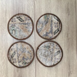 May include: Four circular coasters featuring a woven textile pattern in muted shades of beige, brown, blue, and white. Each coaster is finished with a solid brown stitched border, creating a consistent look across the set of four individual pieces.
