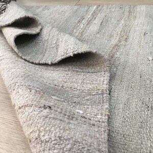 May include: A close-up of a light gray woven rug with a subtle striped pattern. The rug is rolled up and the texture of the weave is visible.
