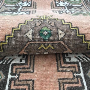 May include: A woven rug featuring a peach-colored background with geometric patterns in dark brown, white, and green. The design includes stepped diamond motifs and intricate borders, showcasing a dense, knotted pile texture throughout the surface.