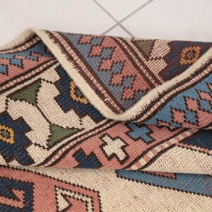 May include: A close-up of a folded rug with a cream background and a geometric pattern in shades of pink, blue, green, and brown.