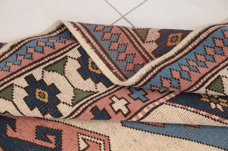 May include: A close-up of a folded rug with a cream background and a geometric pattern in shades of pink, blue, green, and brown.