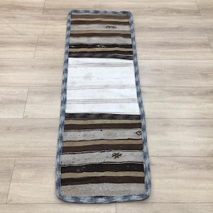 Puede incluir: Una alfombra larga y estrecha con un borde azul y un diseño de patchwork. La alfombra tiene una sección central blanca con rayas marrones y beige a cada lado.