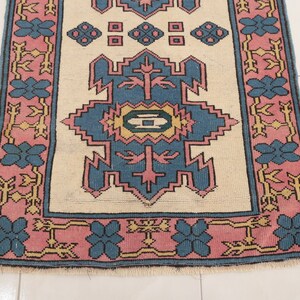 K&ouml;nnte beinhalten: Ein Vintage-Teppich mit cremefarbenem Hintergrund und einem geometrischen Muster in Blau und Rosa. Der Teppich hat einen Fransenrand.