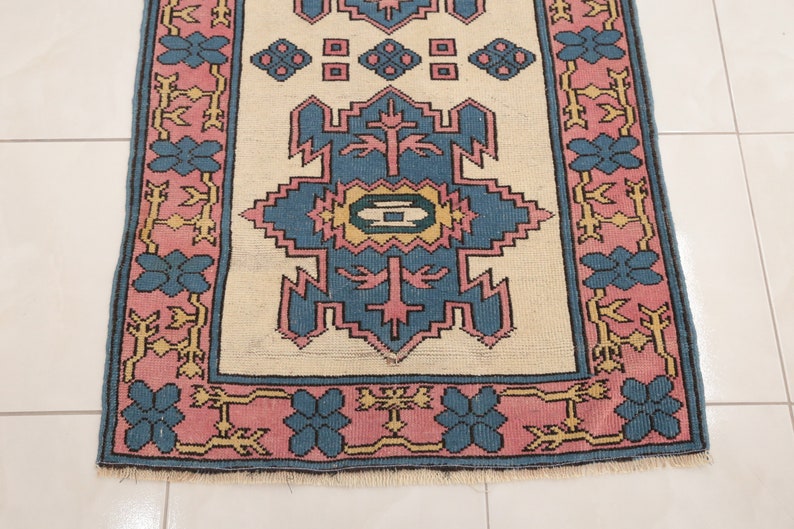 K&ouml;nnte beinhalten: Ein Vintage-Teppich mit cremefarbenem Hintergrund und einem geometrischen Muster in Blau und Rosa. Der Teppich hat einen Fransenrand.