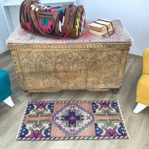 May include: A rectangular wooden chest with intricate floral carvings on the front, topped with a colorful patterned duffel bag and a small stack of books. A small, rectangular woven rug with geometric patterns in pink, purple, and blue lies below.