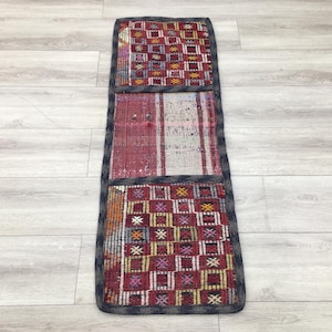 May include: A rectangular, multi-colored rug with a geometric pattern. The rug features a patchwork design with red, white, and orange hues, framed by a dark gray border. The rug is placed on a light-colored wooden floor.