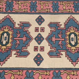 K&ouml;nnte beinhalten: Ein traditioneller Teppich mit cremefarbenem Hintergrund und einem geometrischen Muster in Blau, Rosa und Gelb. Der Teppich hat einen Rand mit rosa und blauen Blumen.
