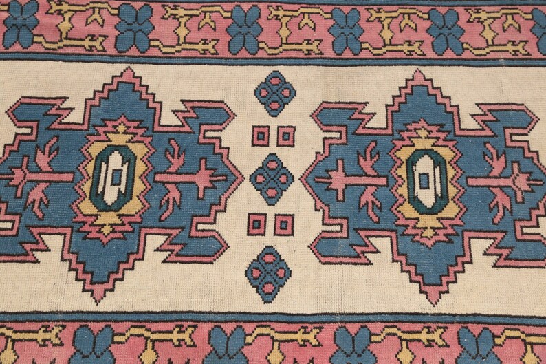 K&ouml;nnte beinhalten: Ein traditioneller Teppich mit cremefarbenem Hintergrund und einem geometrischen Muster in Blau, Rosa und Gelb. Der Teppich hat einen Rand mit rosa und blauen Blumen.