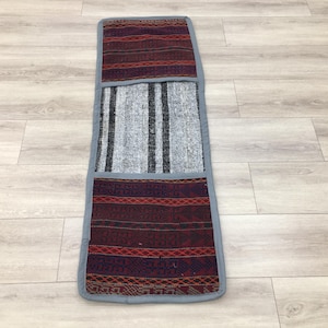 Puede incluir: Una alfombra rectangular larga con un patrón de rayas grises y blancas en el centro. La alfombra tiene un borde estampado en rojo, morado y negro en cada extremo.