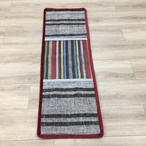 Puede incluir: Una alfombra larga y rectangular con un borde rojo y tres secciones con diferentes patrones. Las secciones superior e inferior son grises con rayas negras, y la sección central es un patrón de rayas de colores.