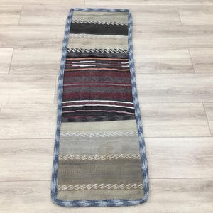Puede incluir: Una alfombra larga y estrecha con un borde azul y un patrón de rayas multicolores. La alfombra tiene una textura tejida y está hecha de fibras naturales.