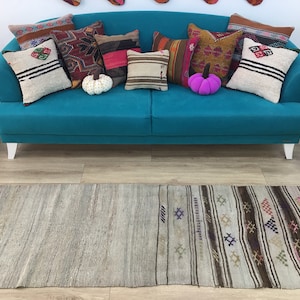 May include: A teal blue sofa with white legs, decorated with colorful patterned throw pillows and a pink pumpkin. A long, woven rug with a striped pattern is in front of the sofa.