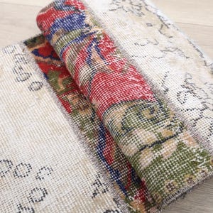 May include: A rectangular rug featuring a patchwork design with sections of off-white fabric displaying faded black floral patterns, contrasted by a central folded section showcasing vibrant red, blue, and green geometric and floral motifs.