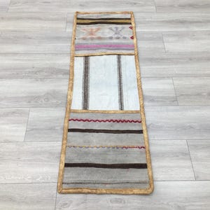 Puede incluir: Una alfombra larga y estrecha con fondo beige y rayas marrones, rojas y blancas. La alfombra tiene un borde marrón con ribete dorado.