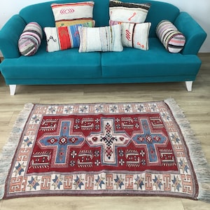 Może przedstawiać: Dywan w kolorze czerwonym, niebieskim i białym z wzorami i frędzlami na krawędziach. Dywan leży na drewnianej podłodze przed turkusową sofą z poduszkami.