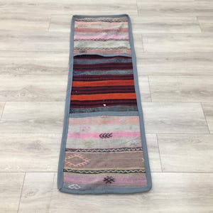 May include: A rectangular, handmade runner rug with a patchwork design. The rug features a variety of colors, including pink, orange, gray, and burgundy. The edges are trimmed with a gray border. The rug is placed on a light-colored wooden floor.