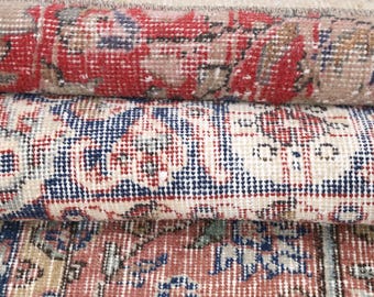 May include: A stack of three rolled rugs featuring intricate woven patterns in shades of red, navy blue, and cream. Each rug displays a distinct geometric and floral motif with a textured surface, showcasing a dense pile and detailed craftsmanship.