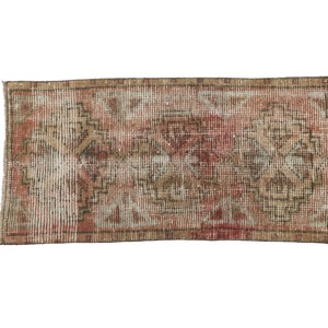 May include: Rectangular rug with a distressed, vintage appearance. The rug features a central design in shades of red, beige, and brown, with a border in a darker brown. The pattern includes geometric and floral elements, creating a textured look.