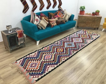 Op de afbeelding: Een lange, rechthoekige loper met een herhalend geometrisch ruitpatroon in tinten rood, blauw, roze, geel en wit, afgewerkt met franjes aan de uiteinden. Het kleed is geweven met een donkere rand en toont een levendig, veelkleurig ontwerp.