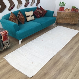Puede incluir: Una alfombra de pasillo blanca con una textura sutil, colocada sobre un suelo de madera marrón claro frente a un sofá azul turquesa con patas blancas. El sofá tiene varios cojines de colores vivos y está situado en un salón.