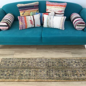 May include: A teal sofa with white legs, featuring two cylindrical striped bolsters and four rectangular throw pillows with multi-colored woven patterns. Below the sofa lies a rectangular rug with a distressed, dark green and gold geometric pattern.