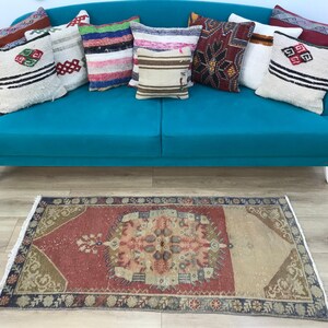 May include: A bright teal sofa with white legs, adorned with nine decorative throw pillows featuring various patterns, colors, and textures. Below the sofa lies a rectangular area rug with a red, beige, and blue floral and geometric pattern.