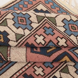 May include: A close-up of a woven rug with a geometric pattern in shades of blue, pink, green, and beige. The rug has a fringe on the edges.