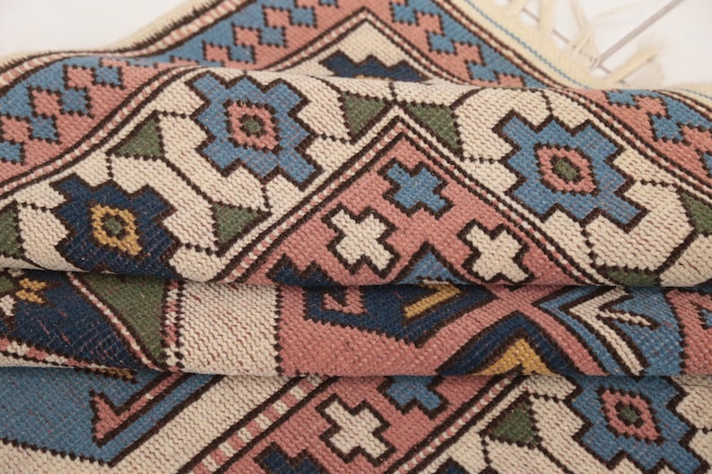 May include: A close-up of a woven rug with a geometric pattern in shades of blue, pink, green, and beige. The rug has a fringe on the edges.