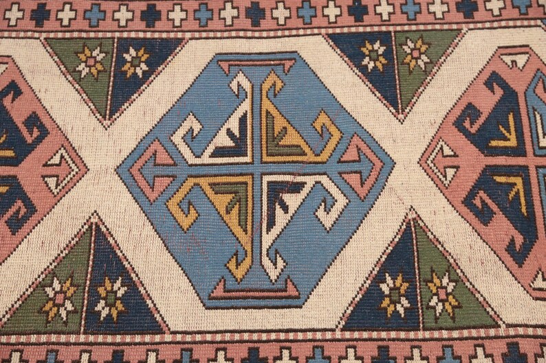 May include: A close-up of a rug with a geometric pattern in shades of blue, pink, and yellow. The pattern is made up of repeating squares and triangles. The rug has a white background.