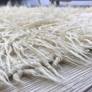 以下が含まれることがあります： 長くふわふわした繊維を持つ白いふわふわの敷物のクローズアップ。敷物は柔らかく、豪華な素材で作られており、自然な漂白されていない外観をしています。