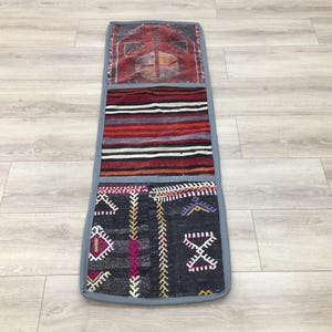 Puede incluir: Una alfombra rectangular larga con tres secciones. La sección superior es roja y blanca con un patrón geométrico. La sección del medio es roja, blanca y negra con rayas horizontales. La sección inferior es negra con un patrón geométrico en tonos rojos, blancos y morados.