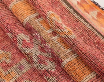 May include: A woven rug featuring a textured pile with a repeating geometric pattern in shades of coral pink, burnt orange, and cream. The fabric shows a dense, knotted construction with visible fiber loops and a fringed edge along the side.