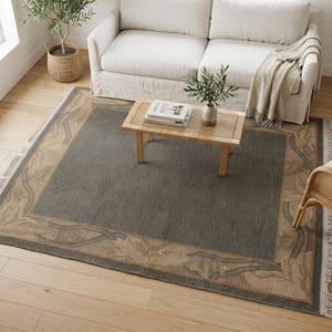 May include: A rectangular area rug featuring a dark charcoal gray center panel surrounded by a wide tan border with a subtle, flowing vine-like pattern. The rug has short, light-colored fringe along the two shorter ends and a woven texture.