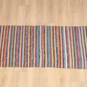 May include: A rectangular rag rug featuring a repeating pattern of vertical stripes in various colors including coral, light blue, dark blue, white, and muted earth tones. The rug has a textured, woven appearance with short fringe at both ends.