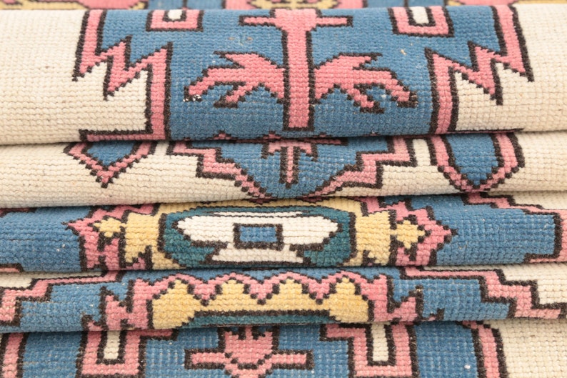 K&ouml;nnte beinhalten: Nahaufnahme eines gefalteten Teppichs mit einem geometrischen Muster in Rosa-, Blau- und Wei&szlig;t&ouml;nen.
