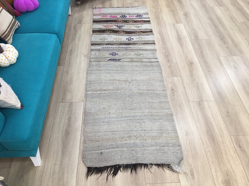 May include: A long, narrow, off-white rug with a fringe on the bottom. The rug has a woven pattern with colorful stripes and geometric designs.
