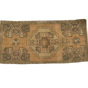May include: A rectangular woven rug featuring a central geometric medallion in shades of tan, brown, and muted green. The design includes symmetrical corner motifs and a repeating pattern of angular lines and shapes across the entire surface.