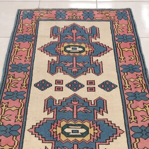 K&ouml;nnte beinhalten: Ein Teppich im Vintage-Stil mit cremefarbenem Hintergrund und einem geometrischen Muster in Blau, Rosa und Gelb. Der Teppich hat einen Rand mit rosa und blauen Blumendesigns.