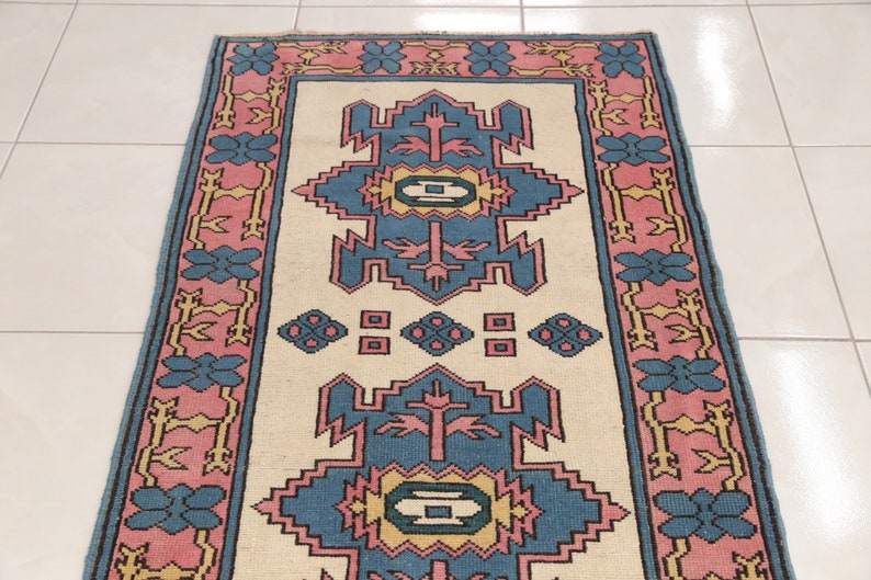 K&ouml;nnte beinhalten: Ein Teppich im Vintage-Stil mit cremefarbenem Hintergrund und einem geometrischen Muster in Blau, Rosa und Gelb. Der Teppich hat einen Rand mit rosa und blauen Blumendesigns.