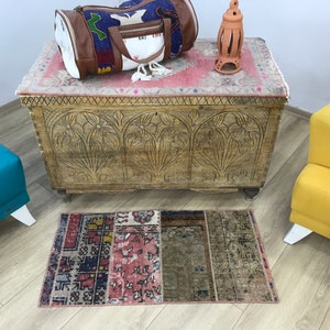 May include: A vintage, hand-woven rug with a patchwork design in shades of red, blue, and brown. The rug is placed on a wooden floor in front of a carved wooden chest.