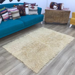 May include: A rectangular, off-white area rug with a long, shaggy pile texture. The rug features a dense, fibrous surface made of soft, light-coloured material, creating a thick and fuzzy appearance that covers the floor in a neutral tone.