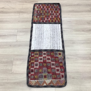 Puede incluir: Una alfombra rectangular larga con una sección central blanca y dos secciones con patrones rojos, amarillos y azules en la parte superior e inferior. La alfombra tiene un borde negro.